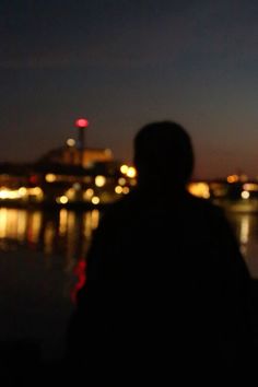 a person standing in front of a body of water at night with the city lights behind them