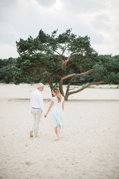 an older couple holding hands and walking in the sand under a tree on a beach