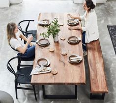 two women sitting at a wooden table with plates and silverware on it, talking to each other