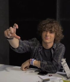 a young man sitting at a table with his hand in the air and pointing to something