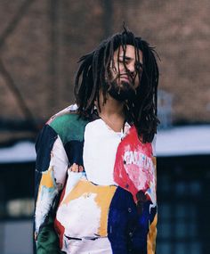 a man with dreadlocks standing in front of a brick building wearing a multicolored shirt