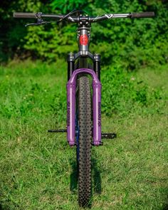 the front end of a purple bike parked on top of a lush green field with trees in the background