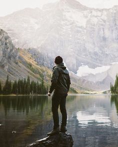 a man standing on top of a rock next to a lake with mountains in the background