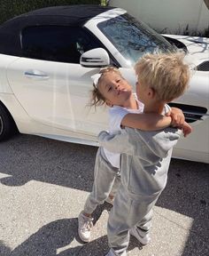 two young children standing in front of a white car with their arms around each other