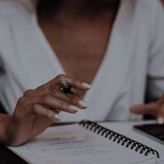 a woman sitting at a table with a notebook and pen in her hand, writing on the paper