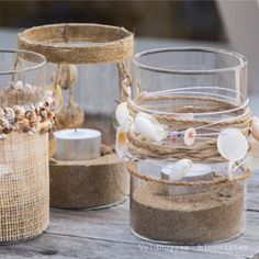 three glass containers filled with sand and seashells on top of a wooden table