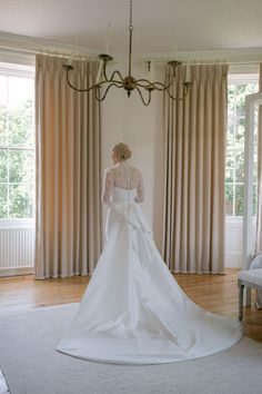 a woman in a white wedding dress is standing near a chandelier and window