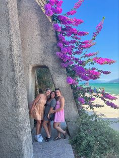 three women are posing for a photo in front of purple flowers on the side of a building