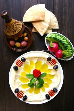 an arrangement of fruits and vegetables on a wooden table with hummus, crackers, olives, and pita chips