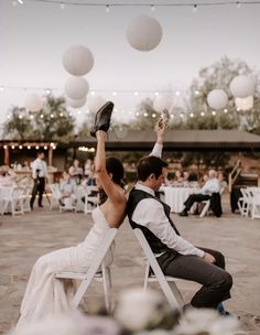 a bride and groom sitting on white folding chairs in front of an outdoor wedding reception