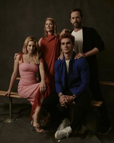 three people sitting on a bench in front of a black background with one person wearing a red dress