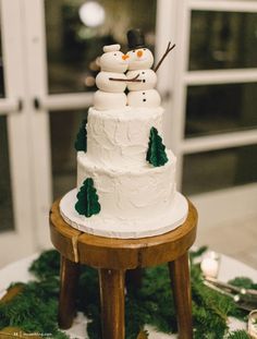 a wedding cake with snowmen on top and evergreen leaves around it, sitting on a small wooden table