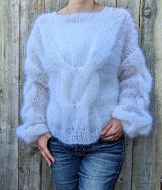 a woman standing in front of a wooden fence wearing a white sweater and blue jeans