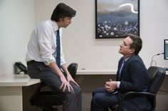 a man in a suit and tie sitting at a desk talking to another man with his hands on his hips