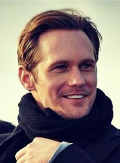 a man wearing a black coat and scarf with his hand on his shoulder smiling at the camera