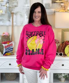 a woman standing in front of a white dresser wearing a pink softball sweatshirt that says softball mom