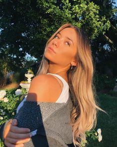 a woman with long blonde hair standing in front of trees and flowers, looking up at the sky