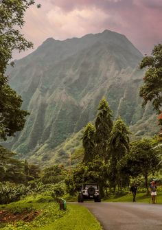 two people are walking down the road in front of a car and some trees with mountains in the background