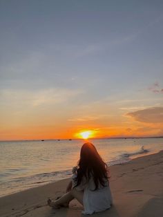 a woman sitting on top of a sandy beach next to the ocean at sun set