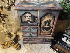 an old fashioned cabinet with two pictures on the front and side, sitting next to other antique items