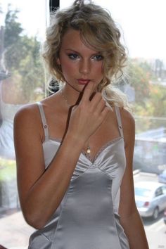 a beautiful young blond woman standing in front of a window with her hand on her chin