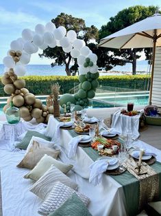 an outdoor table set up for a party with white and green balloons hanging from the ceiling