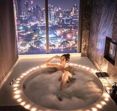 a woman sitting in a bathtub with lights on the side and city lights around it