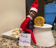 an elf sitting on top of a table next to a bowl of popcorn and a sign that says fresh popcorn