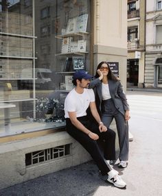 a man and woman sitting on a ledge in front of a store window, both wearing sunglasses