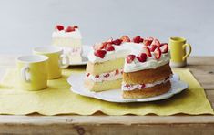 a piece of cake with strawberries on top is sitting on a plate next to two mugs