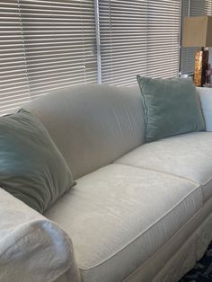 a white couch with pillows on it in front of a window covered by blinds and curtains