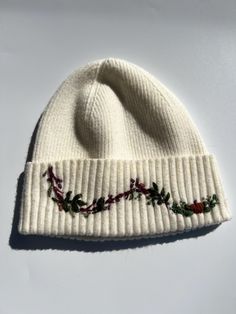 a white beanie with holly embroidered on the front and side, sitting on a white surface