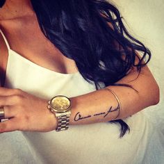 a woman wearing a white tank top and gold bracelets with a wrist tattoo on her arm