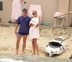 a man and woman standing on the beach next to a small boat with a baby in it