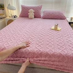 a person reaching for a pink blanket on top of a bed with two pillows and a teddy bear