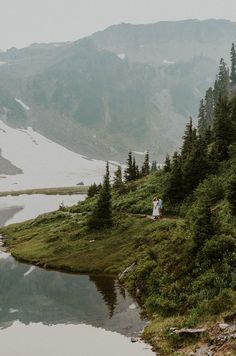 two people are standing on the edge of a mountain overlooking a body of water and trees