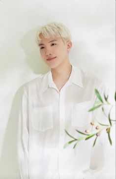 a young man standing next to a white wall with flowers in his hand and wearing a white shirt