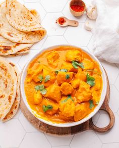 a white bowl filled with chicken curry next to tortillas and pita bread