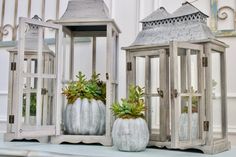 three white lanterns with plants in them sitting on a table next to each other,