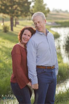 an older man and woman standing next to each other in front of a pond with trees