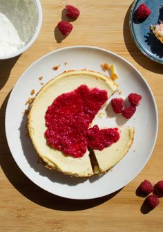 a piece of cheesecake with raspberry topping on a plate next to two bowls of whipped cream and raspberries