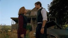 a man and woman standing in front of a house with an old car parked next to them