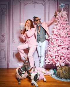 a man and woman posing in front of a pink christmas tree with two dogs on the floor