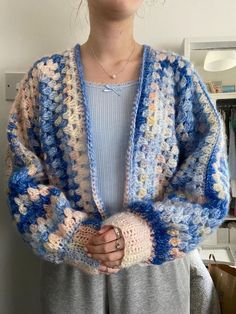 a woman standing in front of a white wall wearing a blue and pink crocheted cardigan