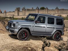 a silver mercedes g - class is parked on the side of a dirt road in front of a castle