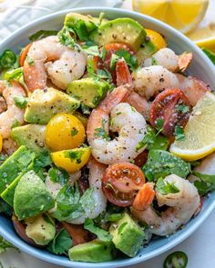 a blue bowl filled with shrimp, avocado and tomato salad next to lemon wedges