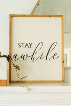 a white framed sign that says stay awhile next to a potted plant on a mantle