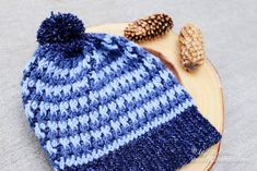 a blue and white knitted hat sitting on top of a wooden plate next to a pine cone