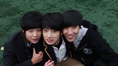 three young men sitting next to each other on top of a grass covered field with one pointing at the camera