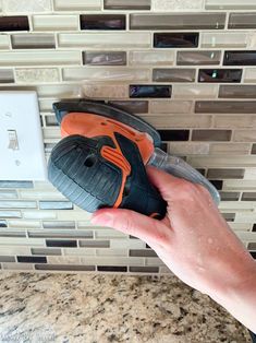 a hand holding an orange and black shoe in front of a wall mounted light switch